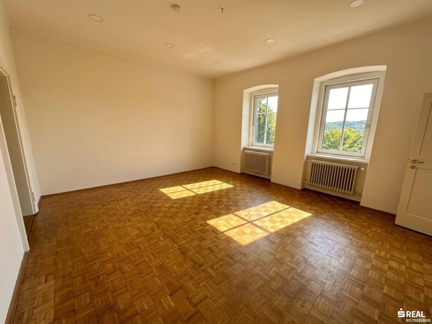 Helles, leeres Zimmer mit Fischgrätparkettboden, weißen Wänden und zwei Fenstern, durch die Sonnenlicht fällt.