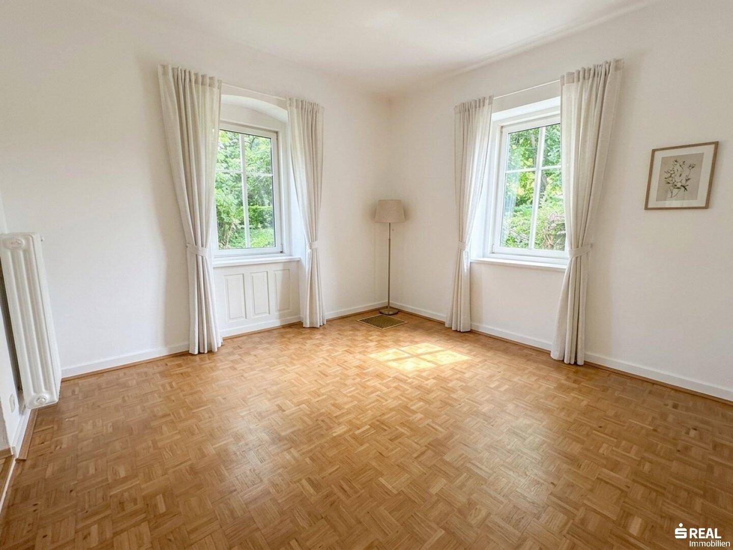 Leeres Zimmer mit Fischgrätparkettboden, weißen Wänden und zwei Fenstern mit hellen Vorhängen.