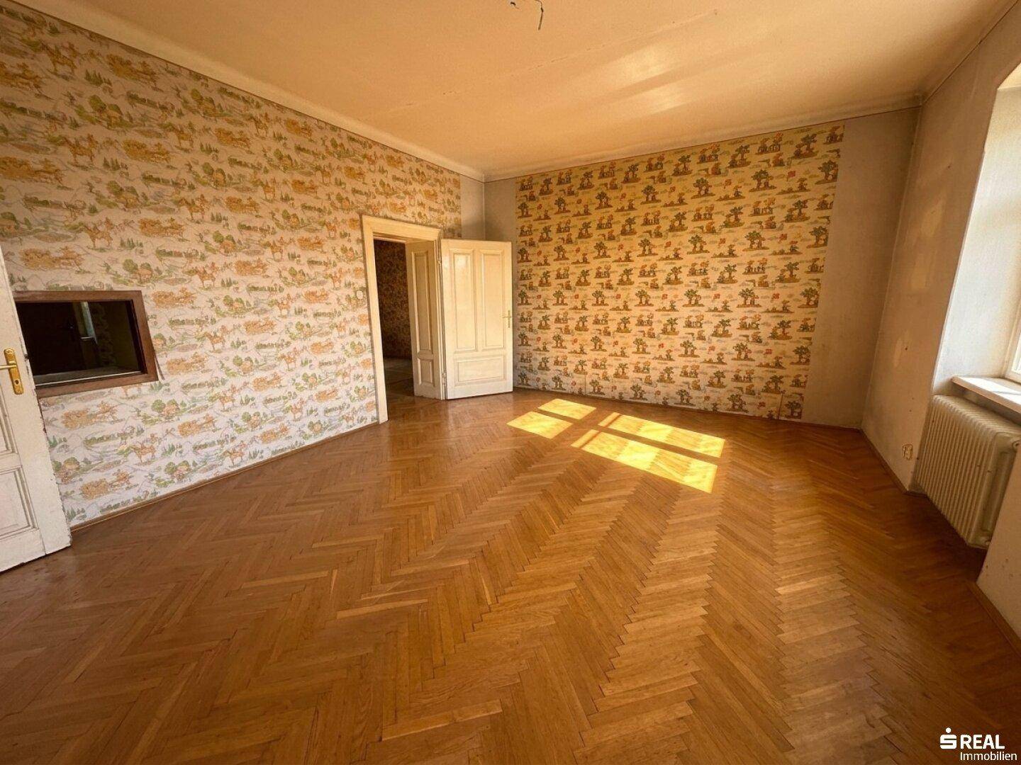 Geräumiges Zimmer mit Fischgrätparkettboden und gemusterter Tapete, durch die Fenster fällt Sonnenlicht.