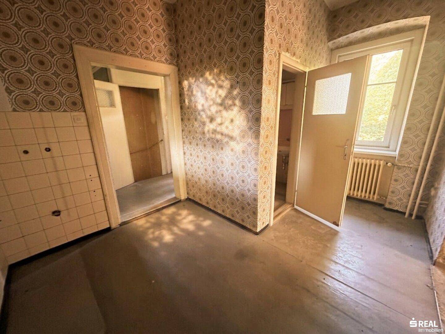 Leerer Raum, möglicherweise eine Küche, mit alten Fliesen, gemusterter Tapete und einem Fenster.