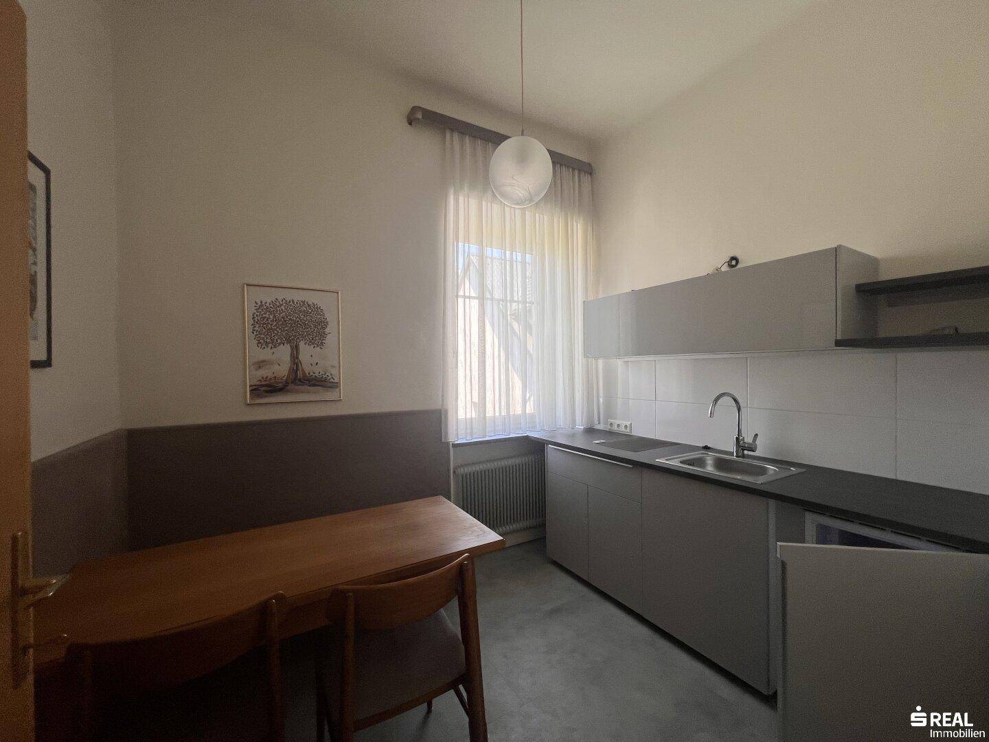 Kleine Küche mit grauen Unterschränken, Spüle und einem Essbereich am Fenster, ideal für Pausen.