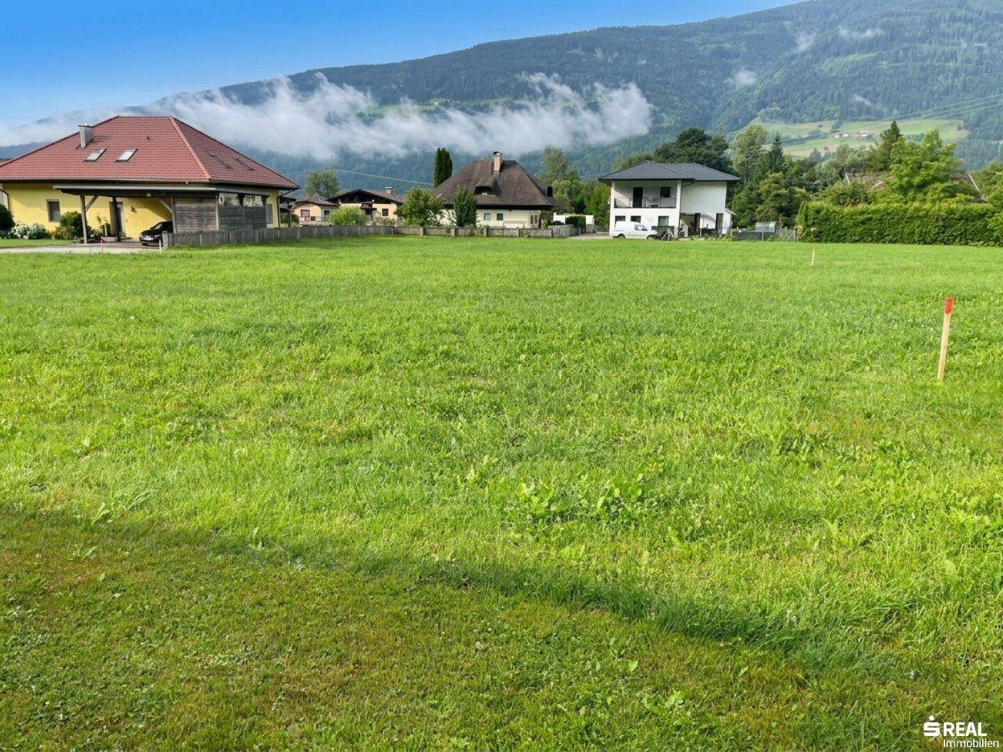 Grüne Wiese mit Blick auf Wohnhäuser und bewaldete Berge unter leicht bewölktem Himmel.