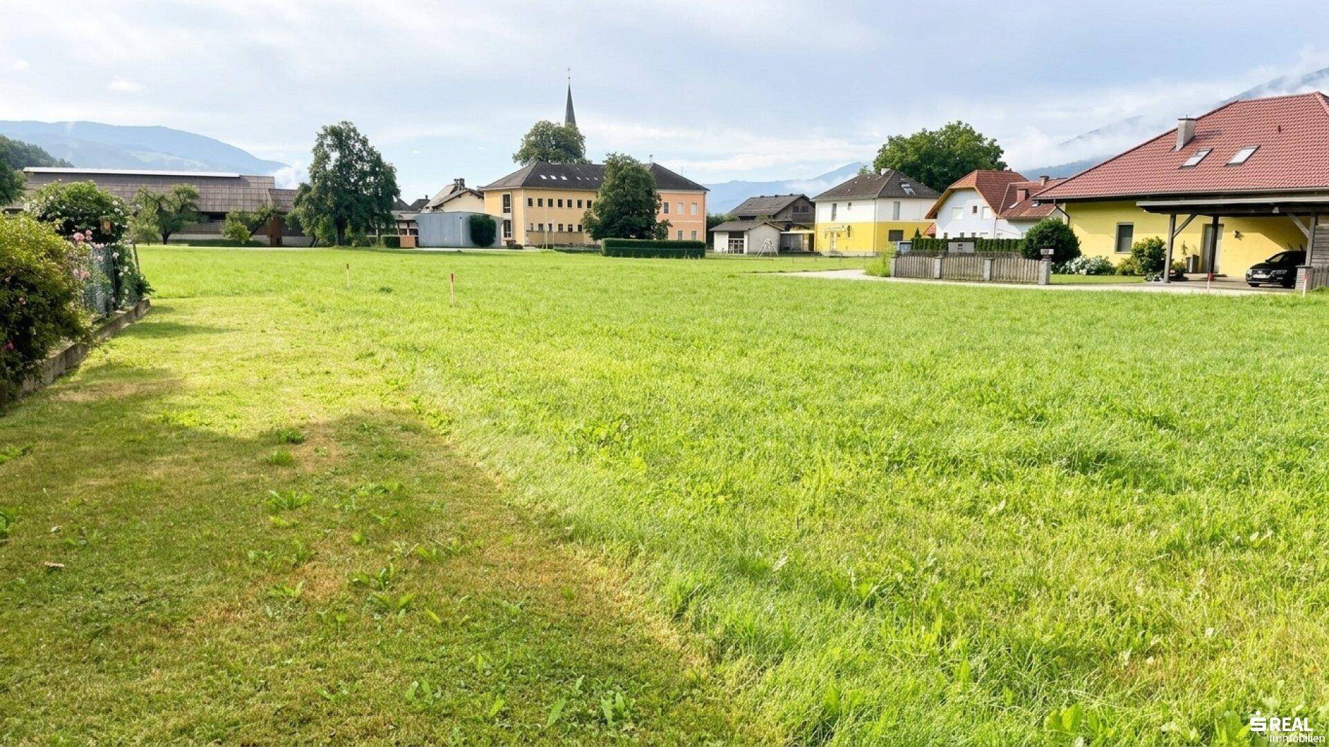 Weitläufige grüne Fläche mit Blick auf ein Dorf, einen Kirchturm und die umliegende Berglandschaft.