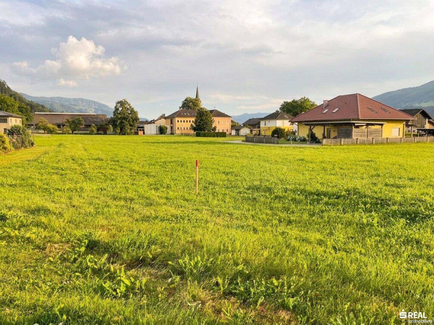 Sonnige grüne Wiese mit Blick auf ein Dorf und die umliegende Berglandschaft am späten Nachmittag.