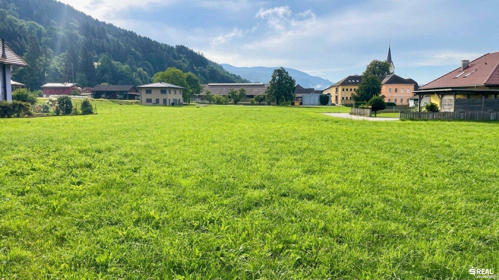 Weitläufige grüne Wiese mit Blick auf umliegende Häuser, Berge und einen Kirchturm unter blauem Himmel.