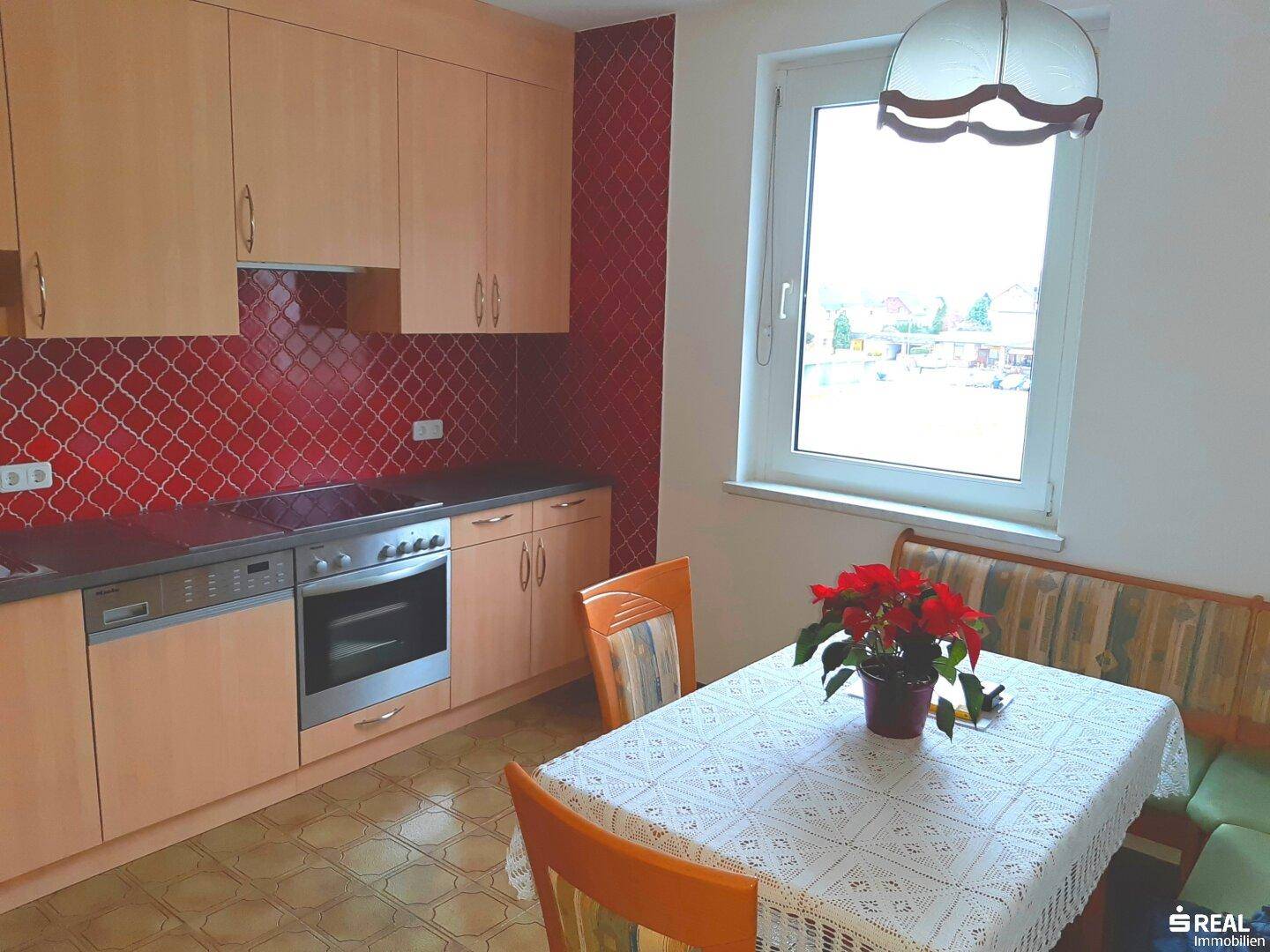 Gemütliche Küche mit hellen Holzschränken, rotem Fliesenspiegel und Essbereich am Fenster.
