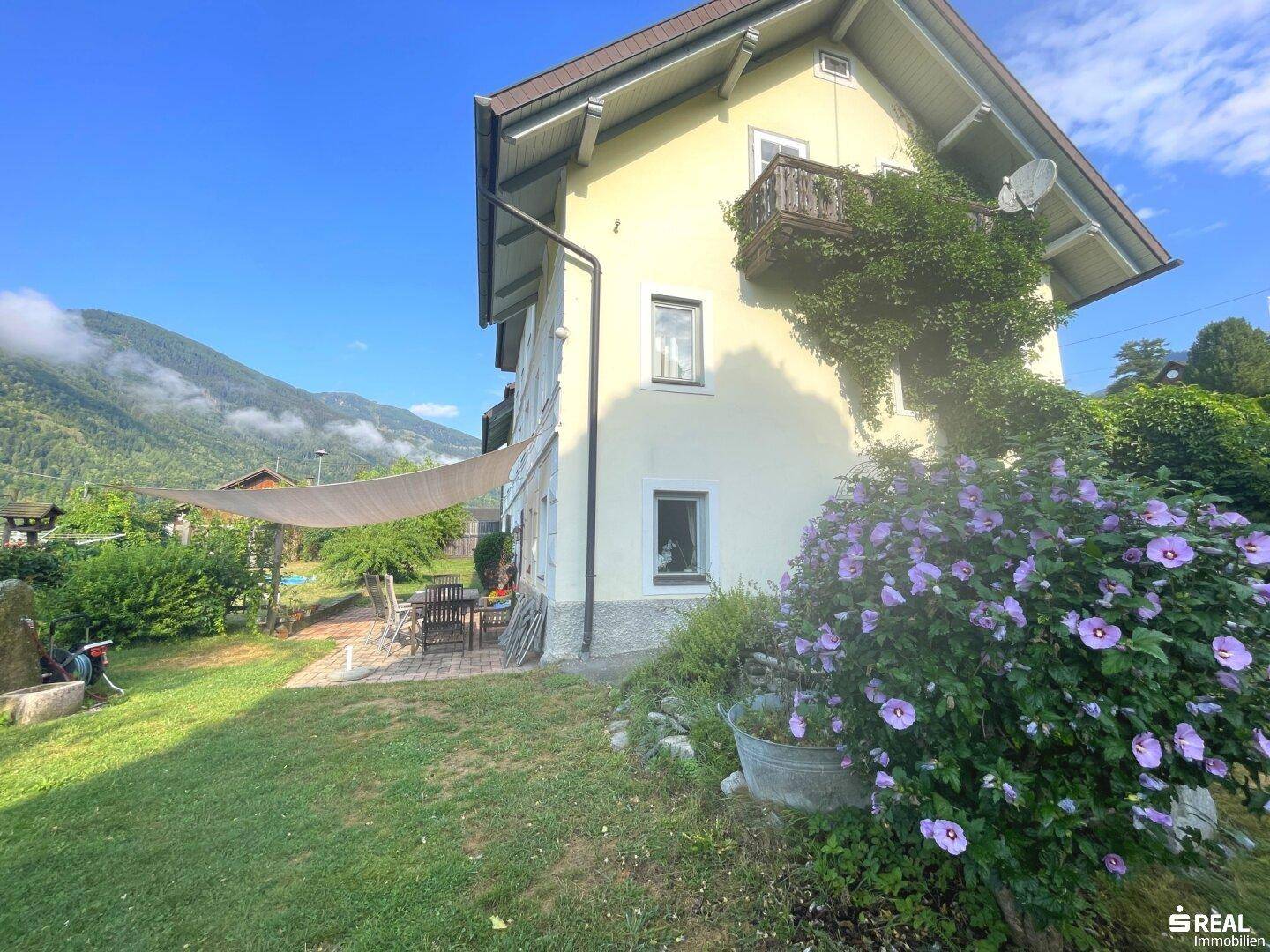 Seitlicher Blick auf das Haus mit Terrasse, Garten und üppiger Vegetation vor Bergkulisse.