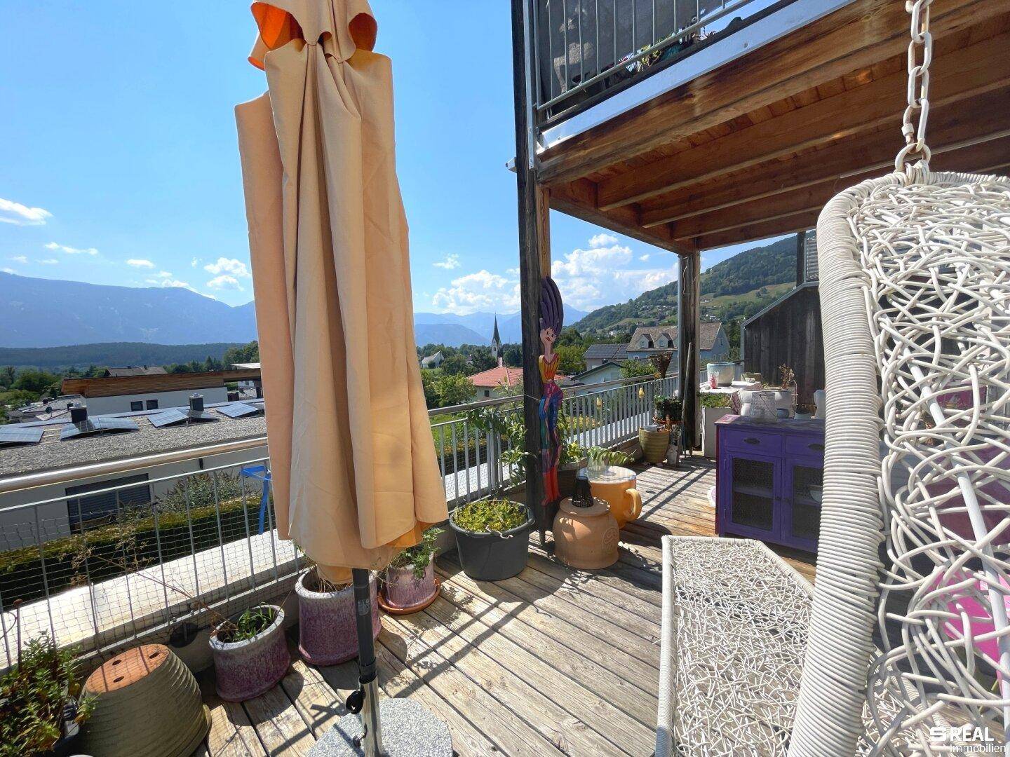Sonnige Terrasse mit Holzboden, Pflanzen, Sitzgelegenheiten und weitem Blick auf die umliegende Berglandschaft.