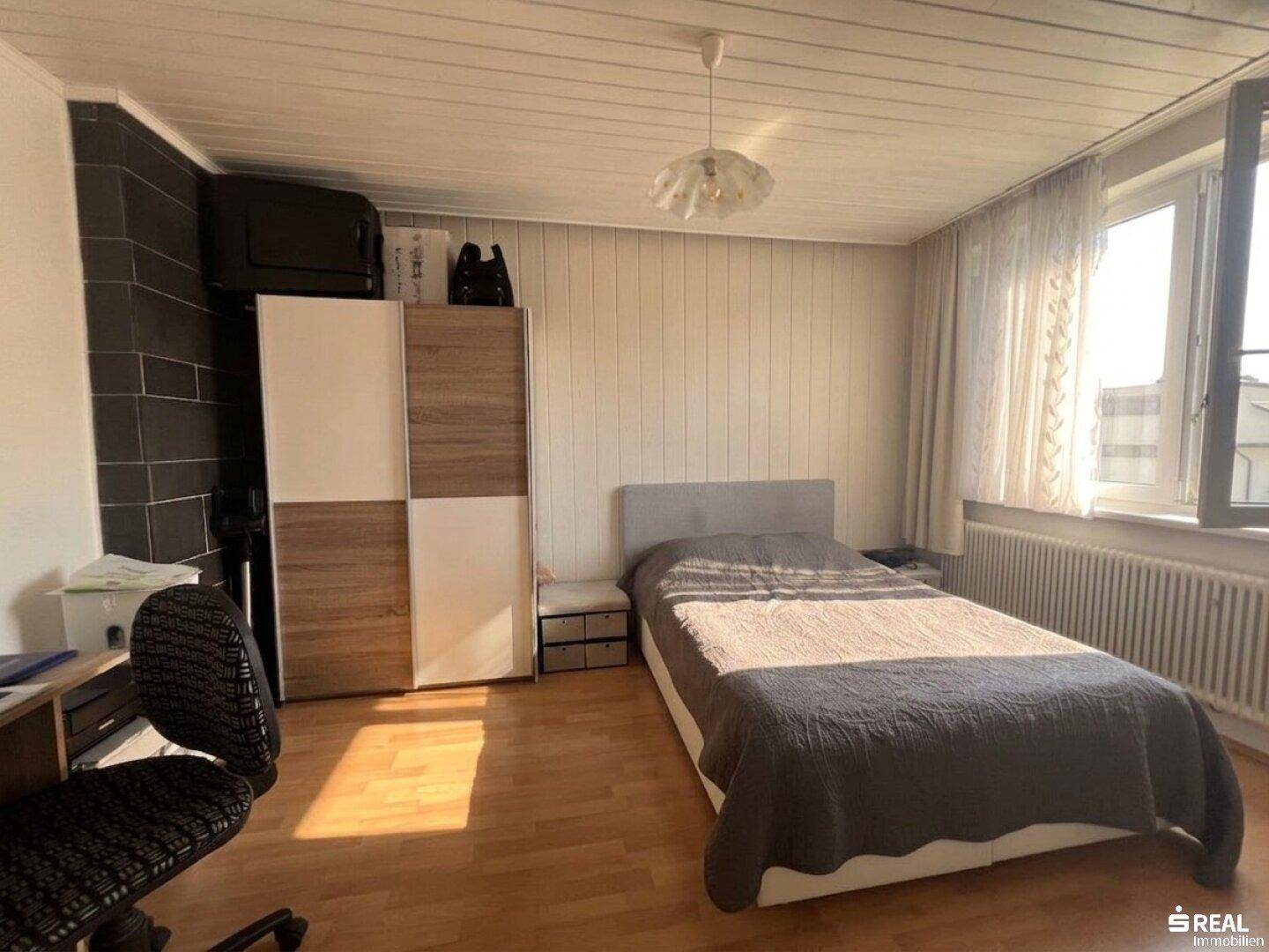 Helles Schlafzimmer mit Einzelbett, Kleiderschrank, Schreibtisch und Holzboden unter einer Holzdecke.