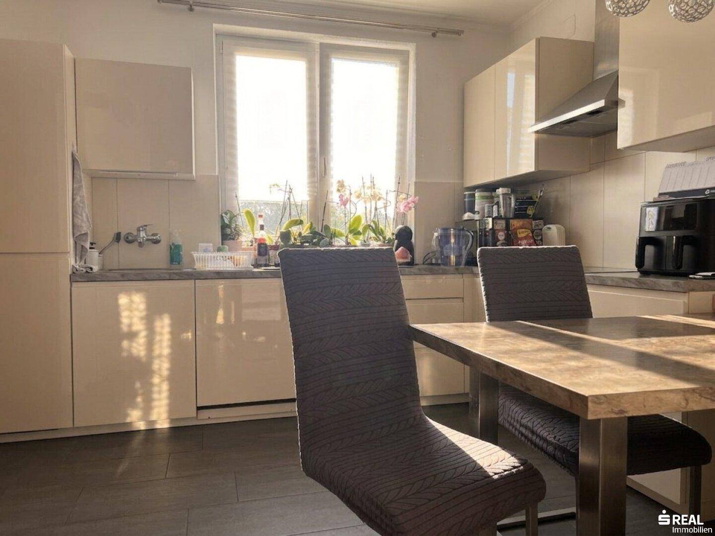 Helle Küche mit modernen beigefarbenen Schränken, dunklem Fliesenboden und Essbereich am Fenster.