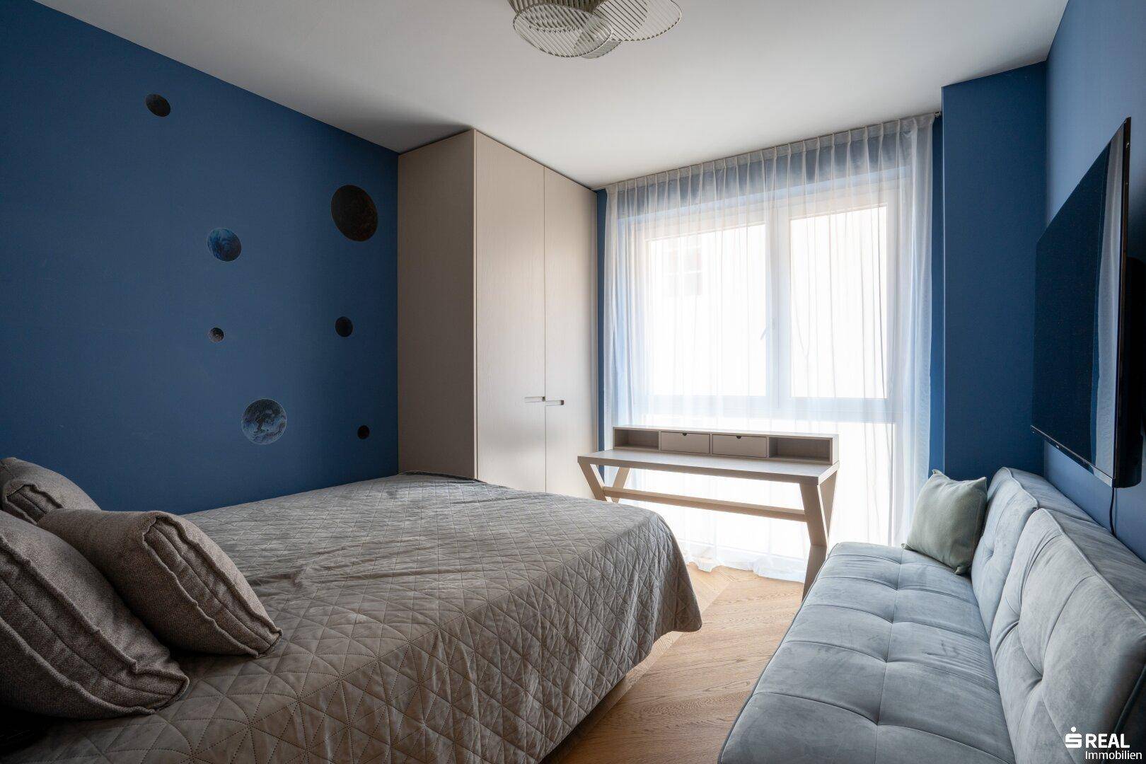 Kinderzimmer mit blau gestrichenen Wänden, Einzelbett und Schreibtisch am Fenster.
