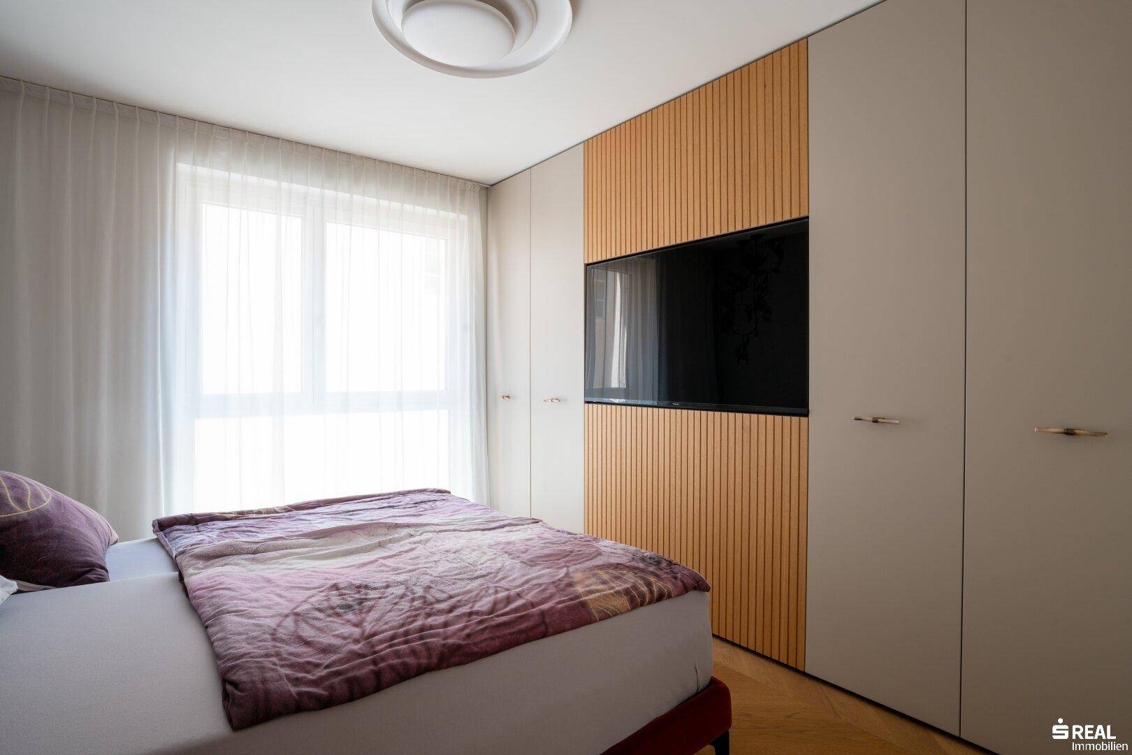 Schlafzimmer mit integriertem TV-Bereich, Einbauschränken und hellem Fenster.