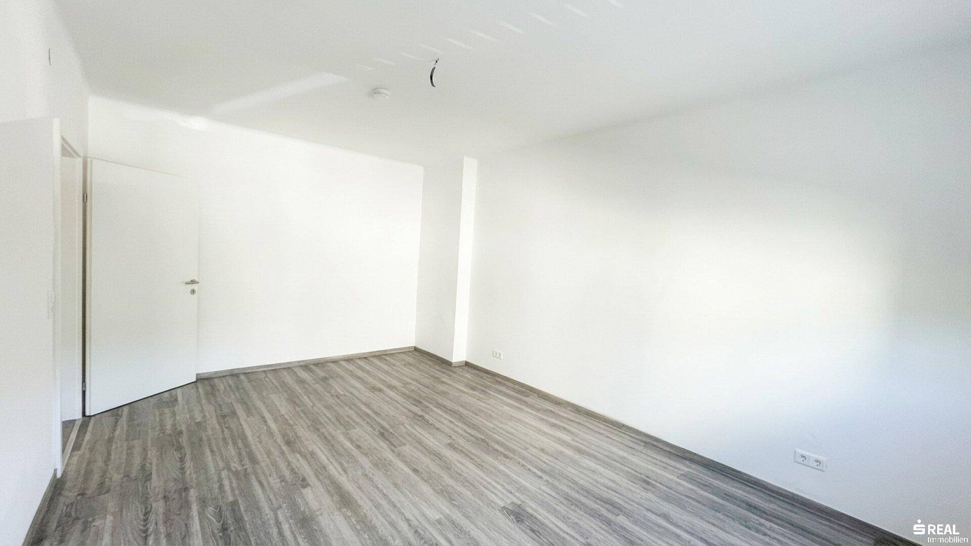 Leerer Raum mit hellem Laminatboden und weiß gestrichenen Wänden.