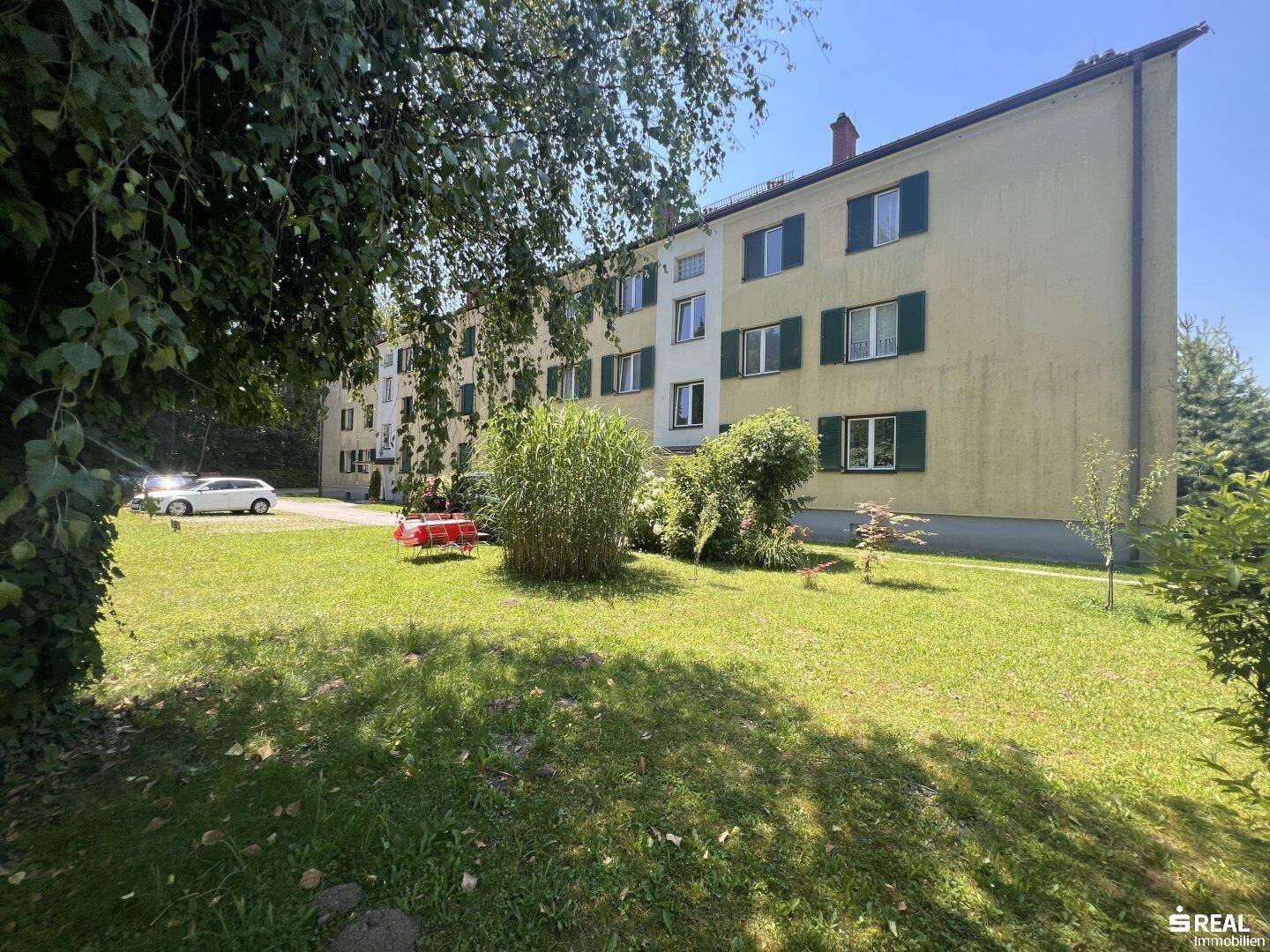 Mehrstöckiges Wohngebäude mit grünen Fensterläden, umgeben von einer gepflegten Rasenfläche und Bäumen.
