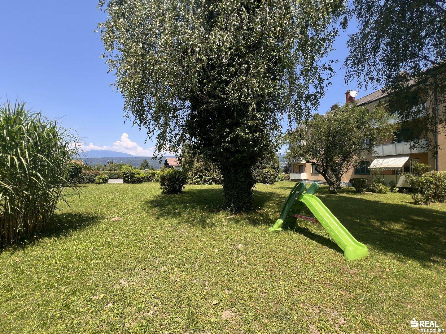 Großer Gemeinschaftsgarten mit Rasenfläche, Bäumen und einem Kinderspielplatz im Freien.