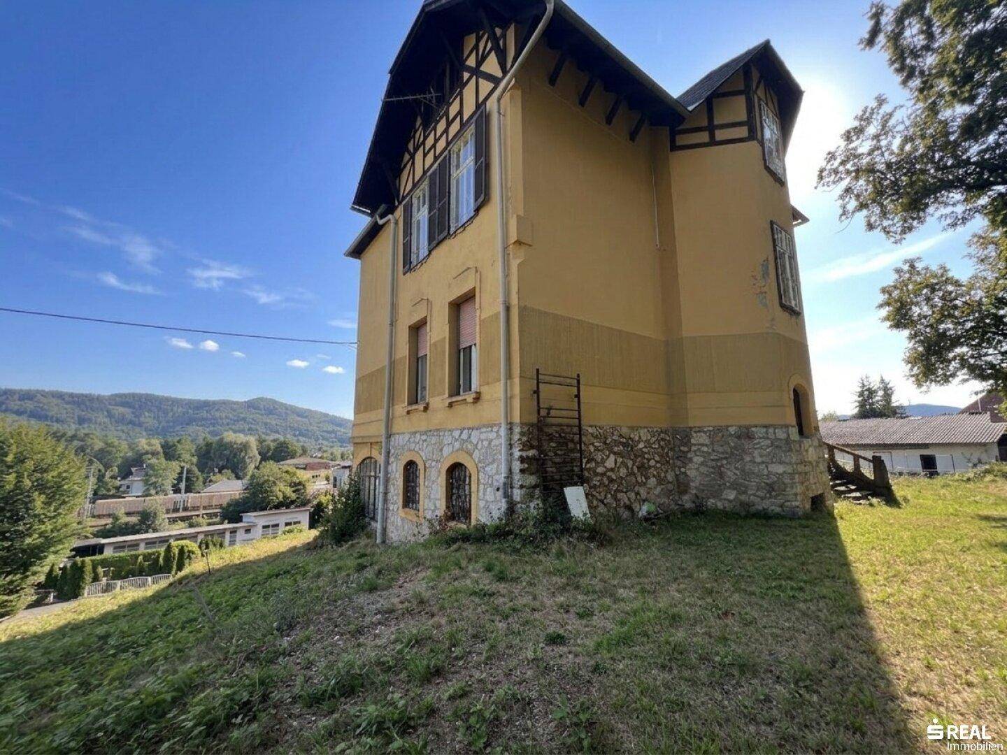 Seitenansicht der historischen Villa mit gelber Fassade, Steinsockel und Blick auf die Umgebung.