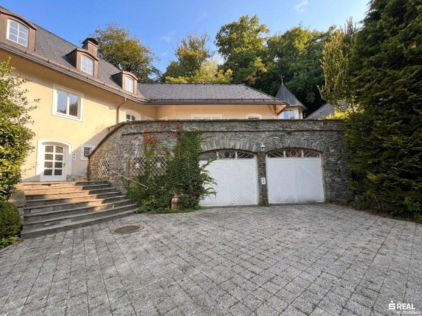 Elegante Villa mit Natursteinmauer und zwei Garagentoren, umgeben von gepflegter Auffahrt.