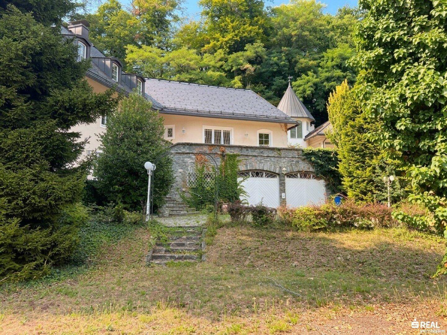 Villa mit Natursteinmauer und Garagenzugang, umgeben von altem Baumbestand und üppiger Vegetation.
