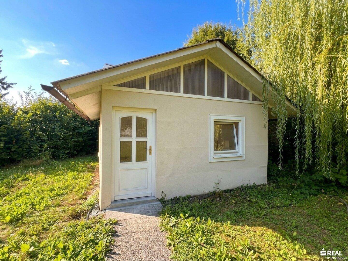 Kleines Gartenhaus mit Giebeldach und weißer Tür, umgeben von grünem Rasen und Bäumen.