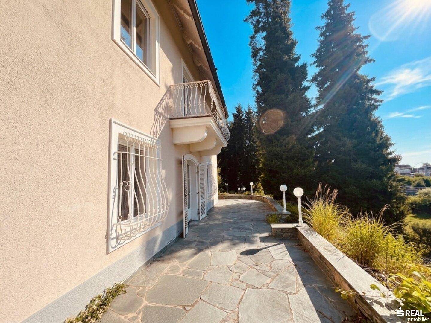Steinplattenweg entlang der Villa mit dekorativen Lampen und Blick auf die umliegende Natur.
