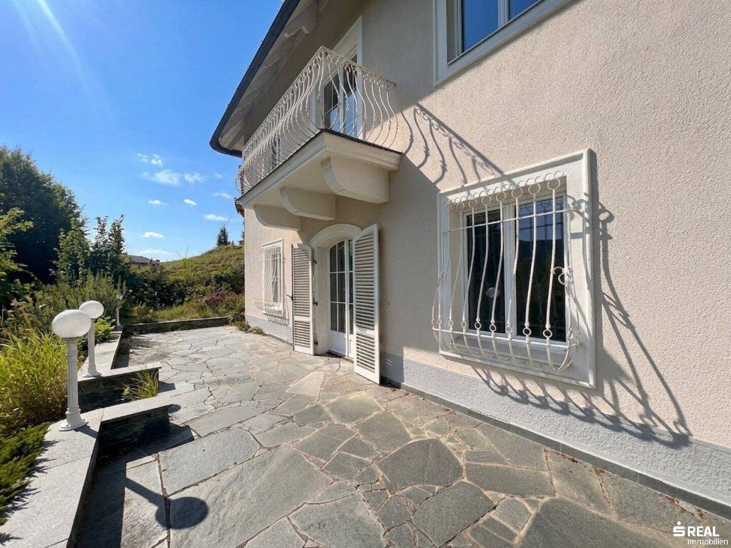 Sonniger Steinplattenweg entlang der Villa mit schmiedeeisernem Geländer und Blick ins Grüne.