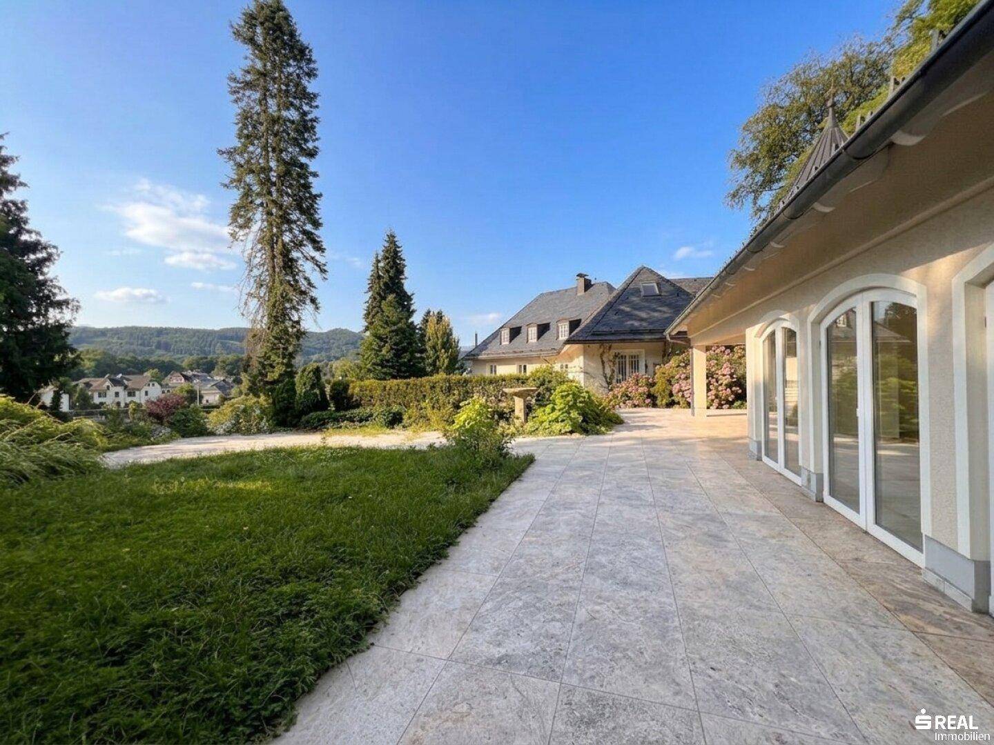 Weitläufige Terrasse mit hellem Steinboden und angrenzendem Rasen, mit Blick auf die Landschaft.