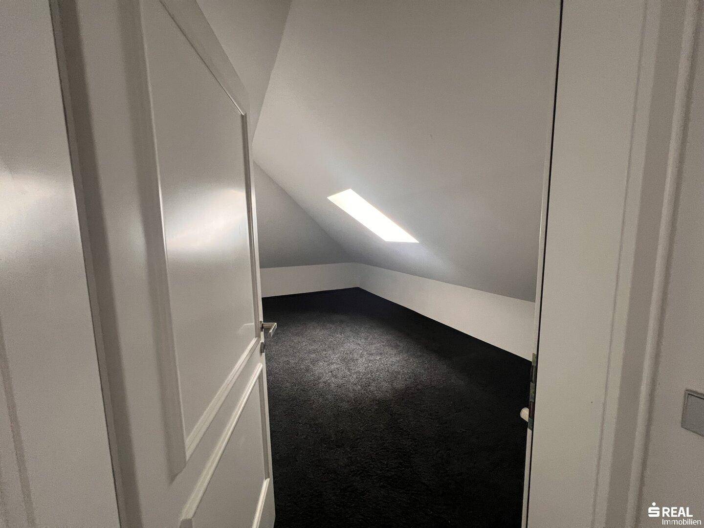 Dachbodenraum mit dunklem Teppichboden und Dachfenster, bietet Potenzial für Ausbau.