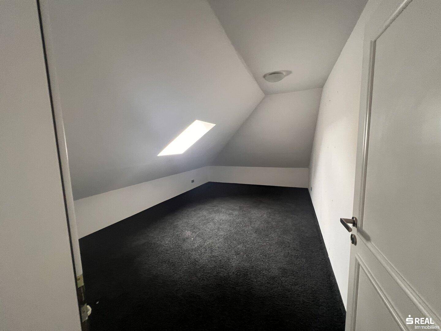 Heller Dachbodenraum mit Dachfenster und dunklem Teppichboden, bereit für individuelle Gestaltung.