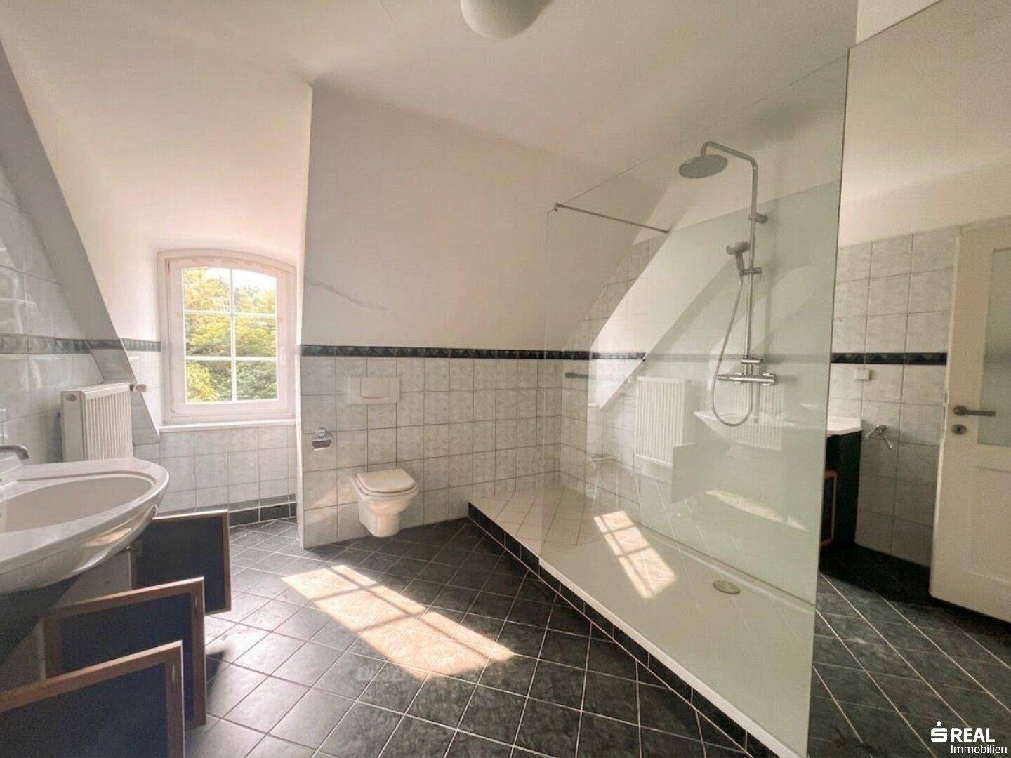 Geräumiges Badezimmer mit begehbarer Dusche, Fenster und dunklen Bodenfliesen.