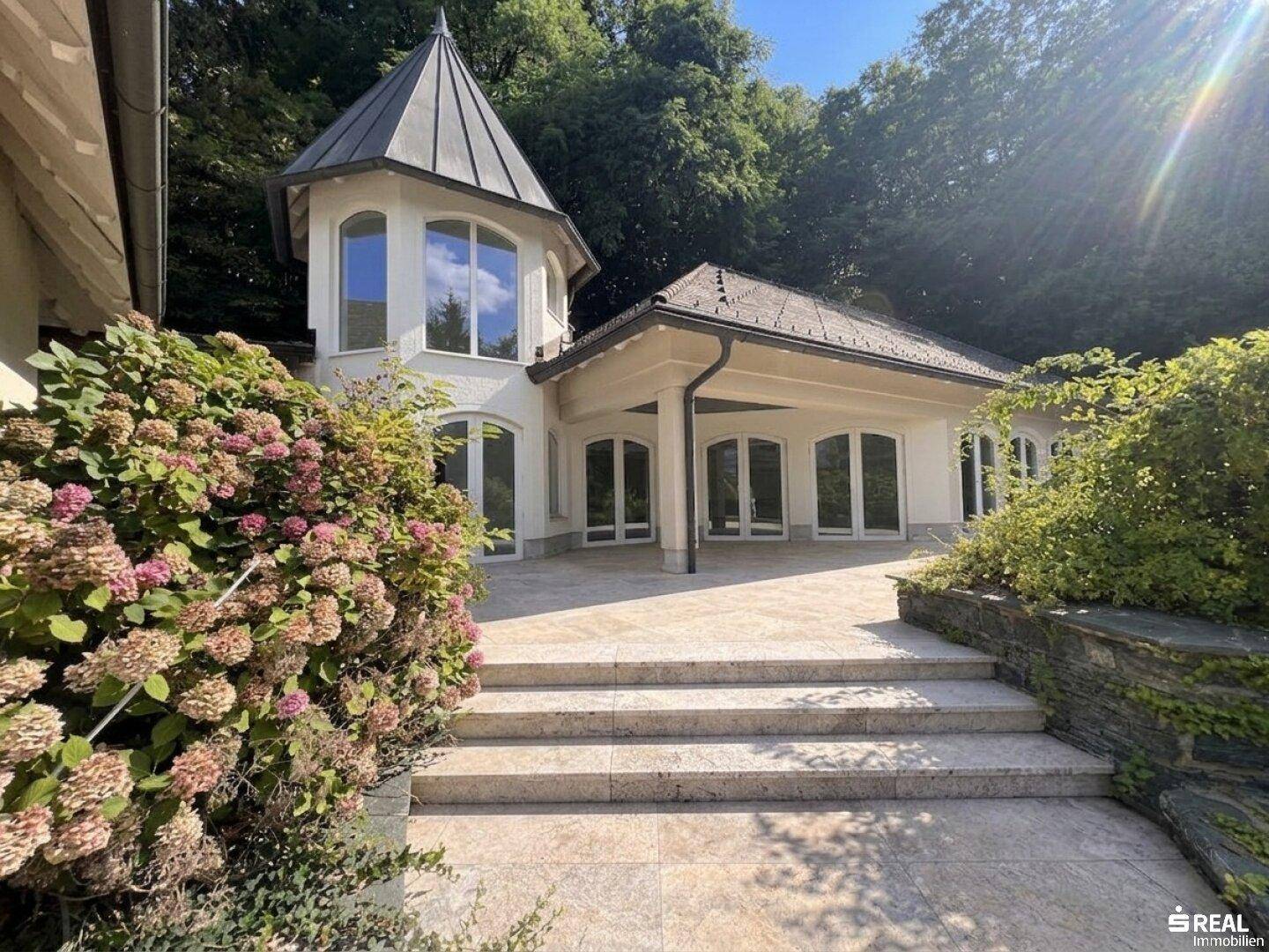Elegante Villa mit Turm und großer Steinterrasse, umgeben von üppigem Grün.