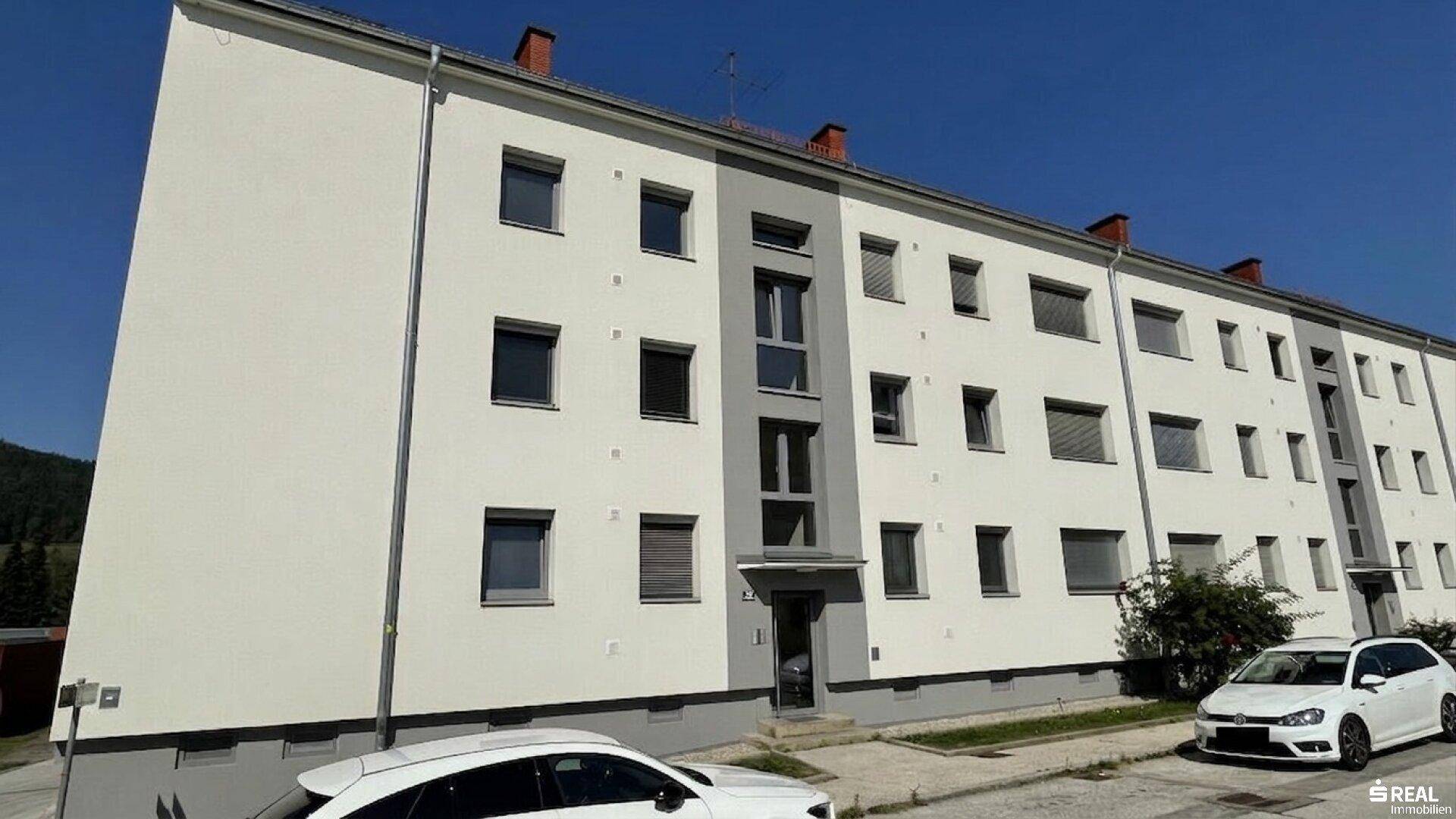 Mehrfamilienhaus mit weißer Fassade und grauen Akzenten, umgeben von Grün und mit Bergblick.