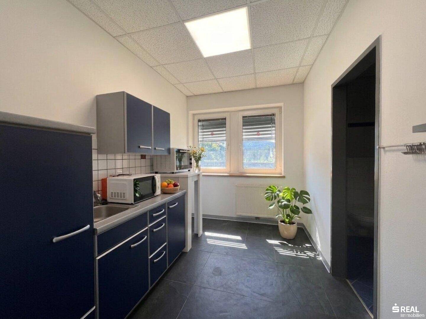 Kleine Küchenzeile mit blauen Schränken, Spüle, Mikrowelle und Fenster mit Jalousien.