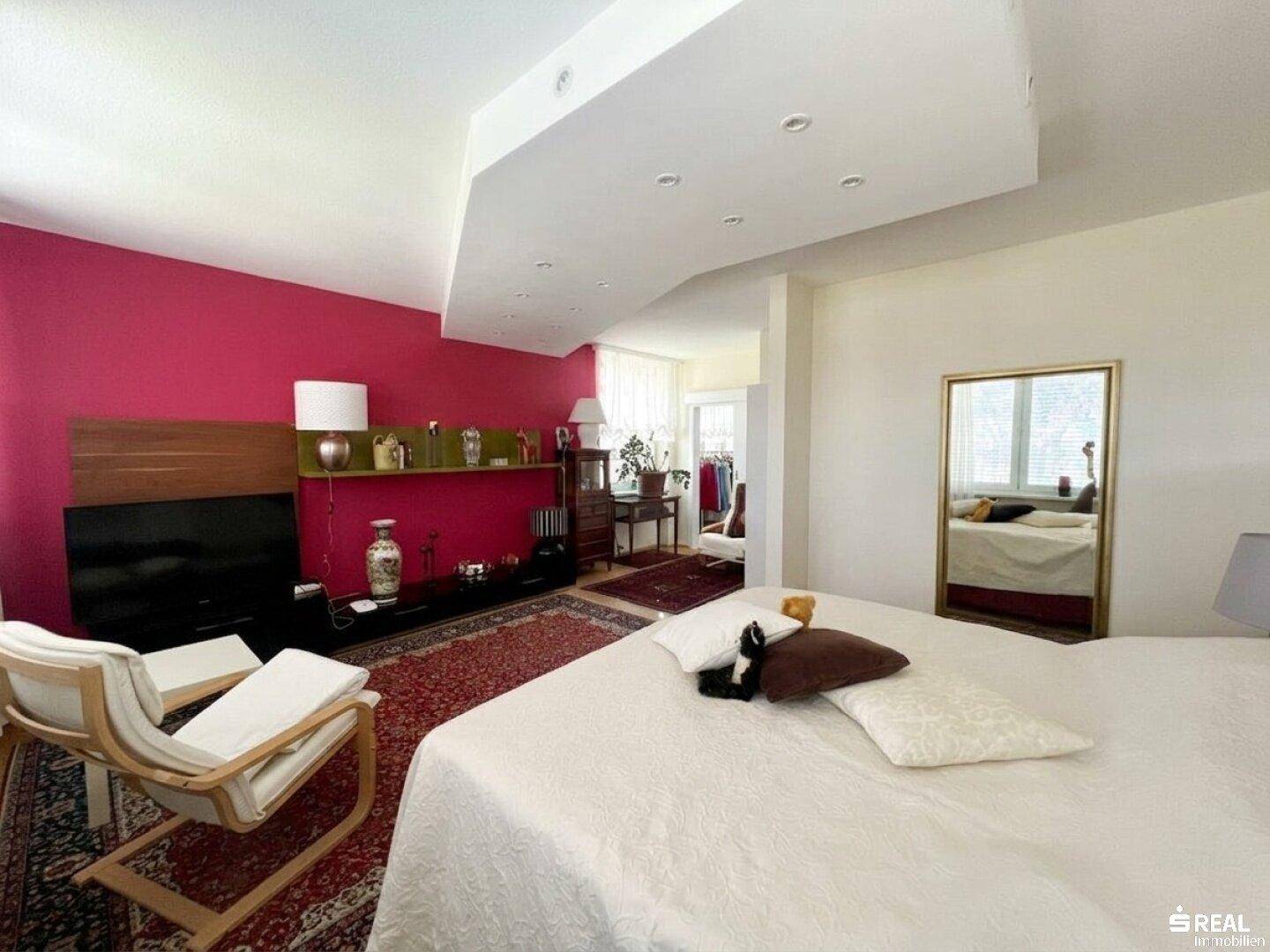 Helles Schlafzimmer mit großem Bett, roter Wand und separatem Arbeitsbereich am Fenster.