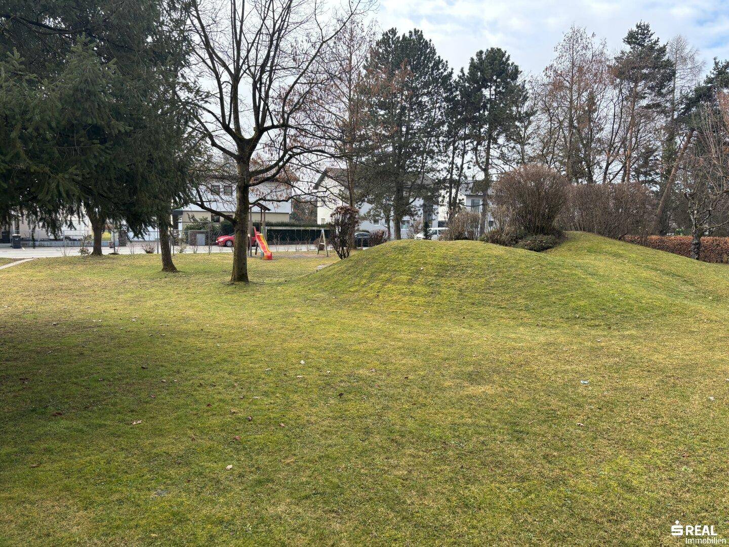 Große Gemeinschaftsgrünfläche mit Rasen, Bäumen und einem kleinen Spielplatz.
