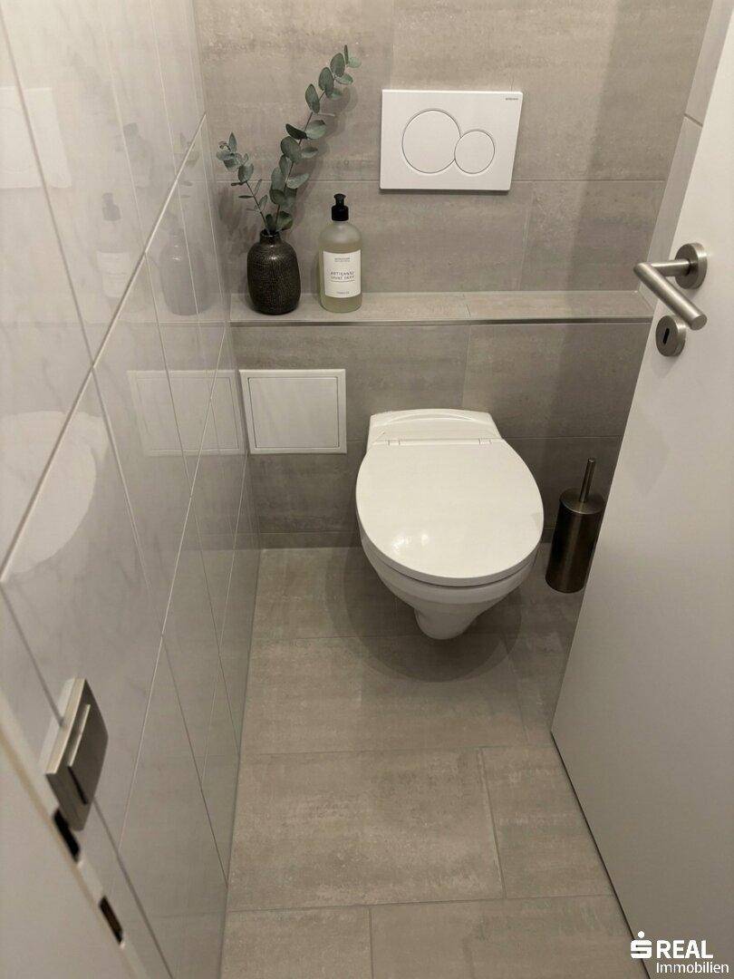 Modernes Gäste-WC mit wandhängender Toilette und grauen Fliesen an Wänden und Boden.