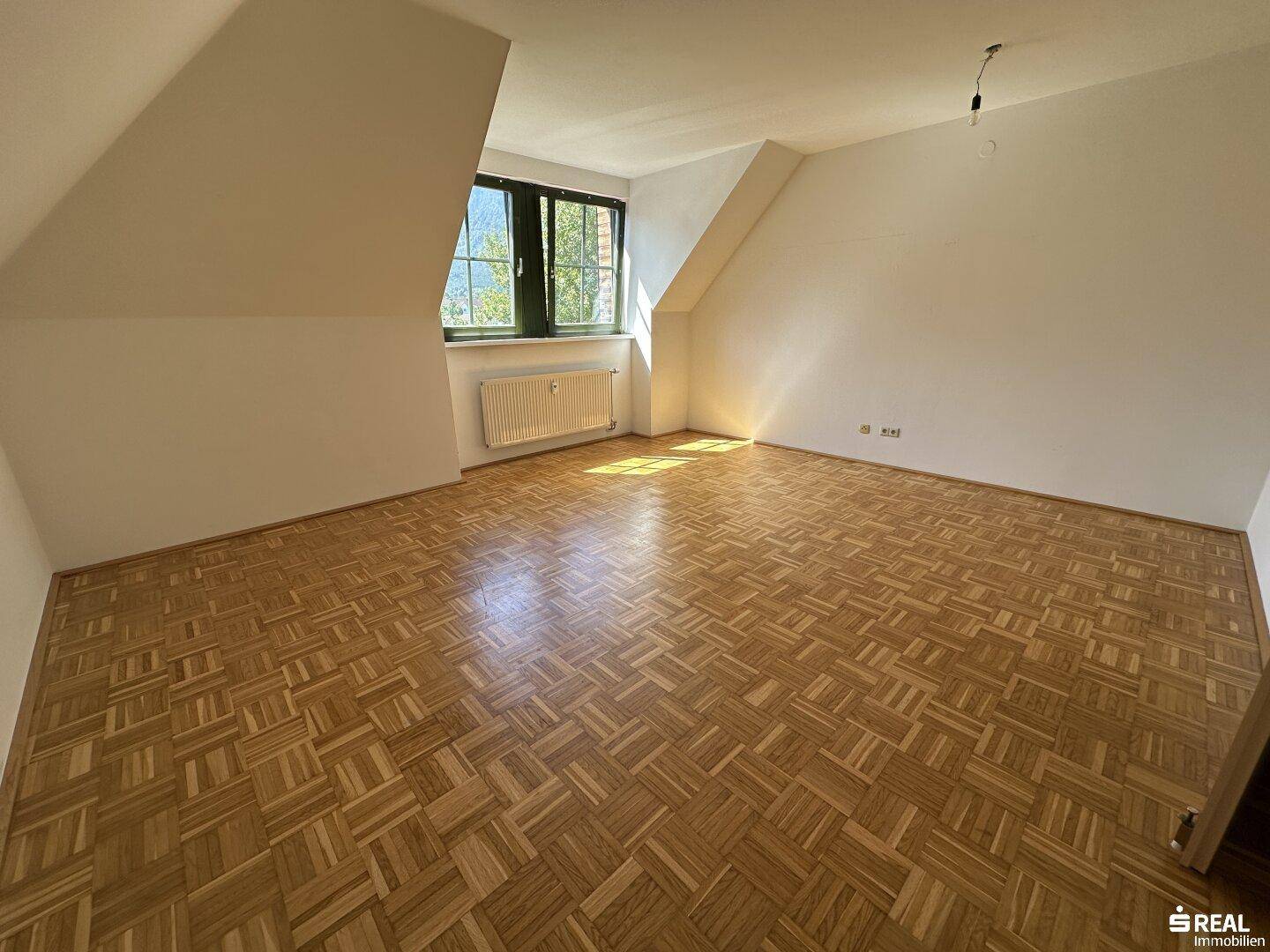 Helles Zimmer im Dachgeschoss mit Parkettboden, schrägen Wänden und einem Fenster mit Blick ins Grüne.