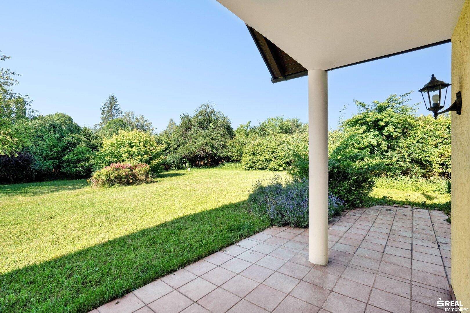 Überdachte Terrasse mit Fliesenboden und direktem Zugang zum weitläufigen, grünen Garten.