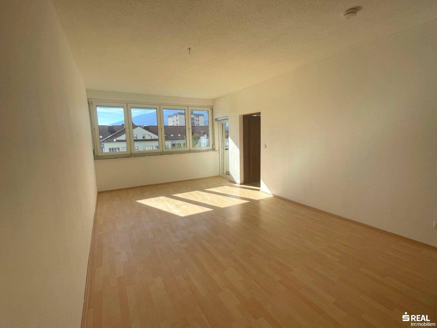 Geräumiges, helles Wohnzimmer mit Holzboden und großen Fenstern, die viel Tageslicht hereinlassen.
