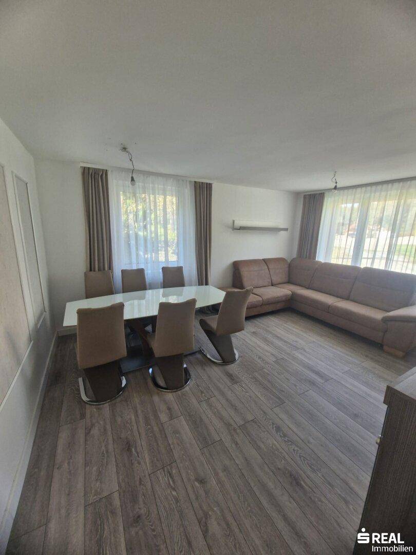 Geräumiger Wohn-Essbereich mit großem Esstisch, bequemer Couch und viel Tageslicht durch die Fenster.