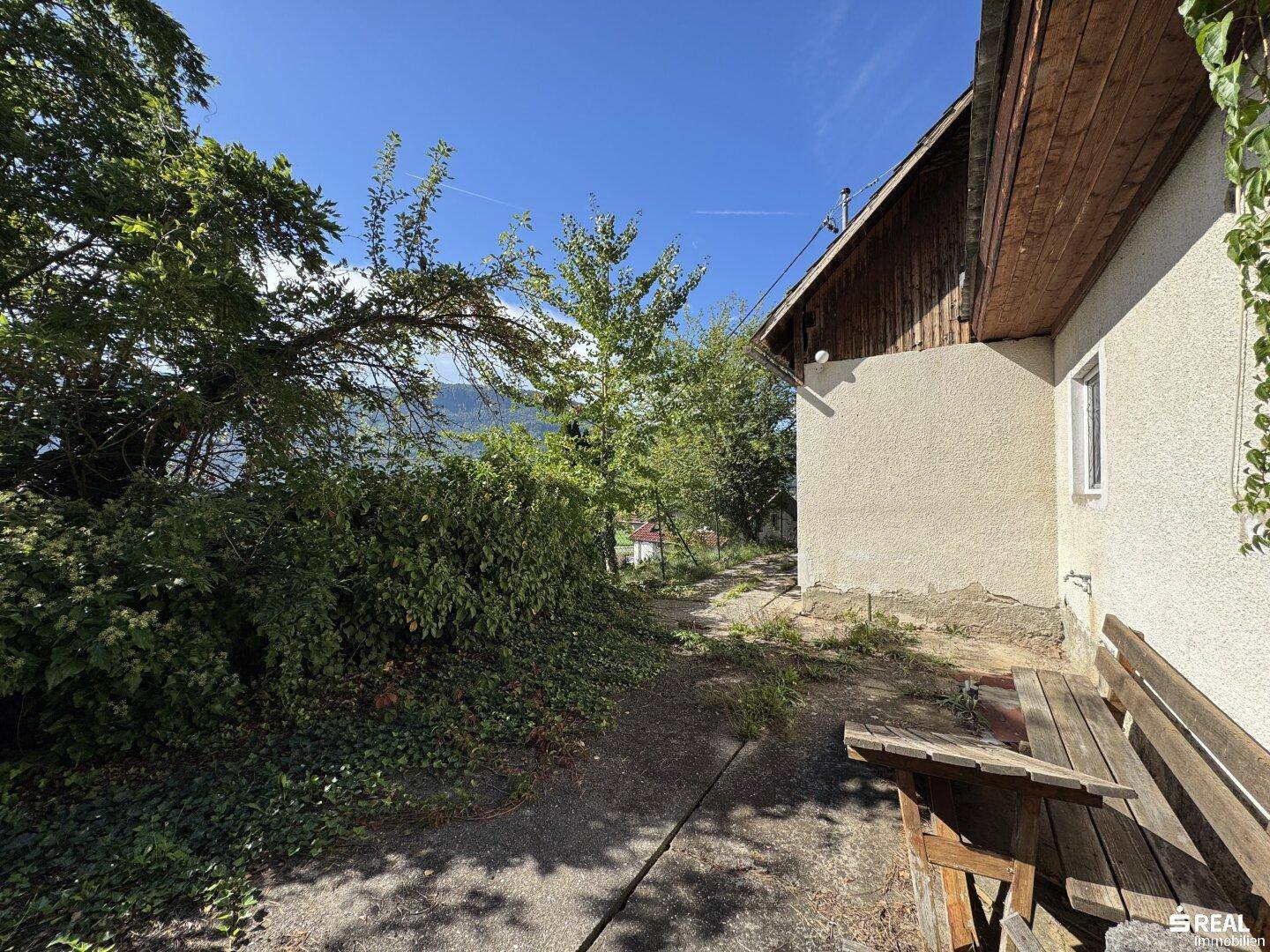 Außenbereich des Hauses mit einer kleinen gepflasterten Fläche, Holzbank und üppiger Vegetation.