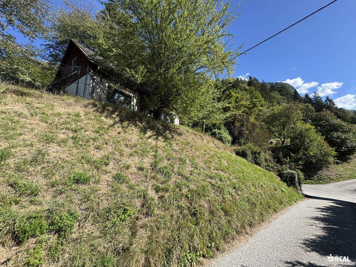 Haus auf einem grasbewachsenen Hang neben einem Feldweg, umgeben von Bäumen und Bergen.