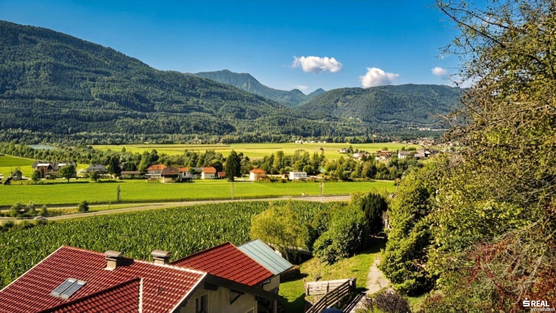 Weitblick über eine ländliche Landschaft mit roten Dächern, grünen Feldern und bewaldeten Bergen.