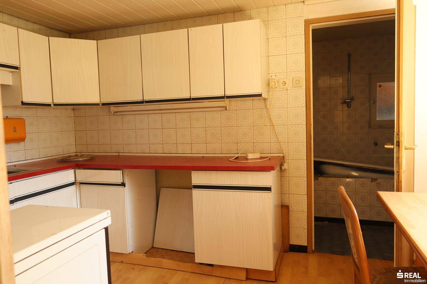 Küche mit weißen Schränken, roten Arbeitsplatten und gefliesten Wänden, mit Zugang zum Badezimmer.