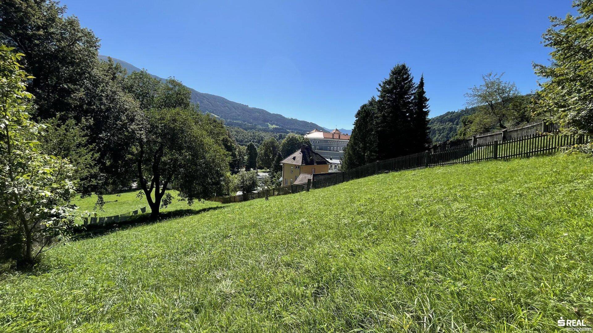 Sonnige grüne Hügellandschaft mit Blick auf entfernte Gebäude und die majestätischen Berge.