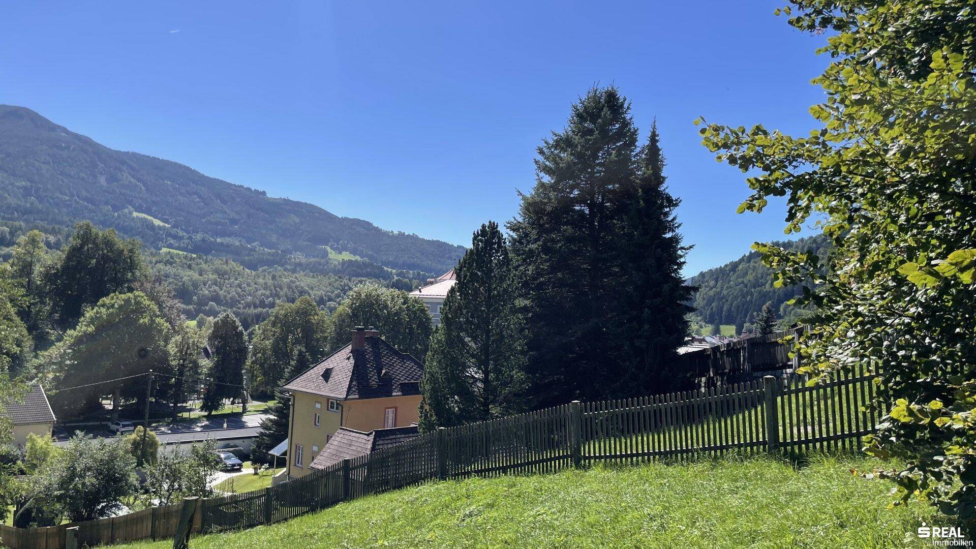 Panoramablick von einer grünen Anhöhe auf ein Wohnhaus und die umliegende Natur mit Bergen.