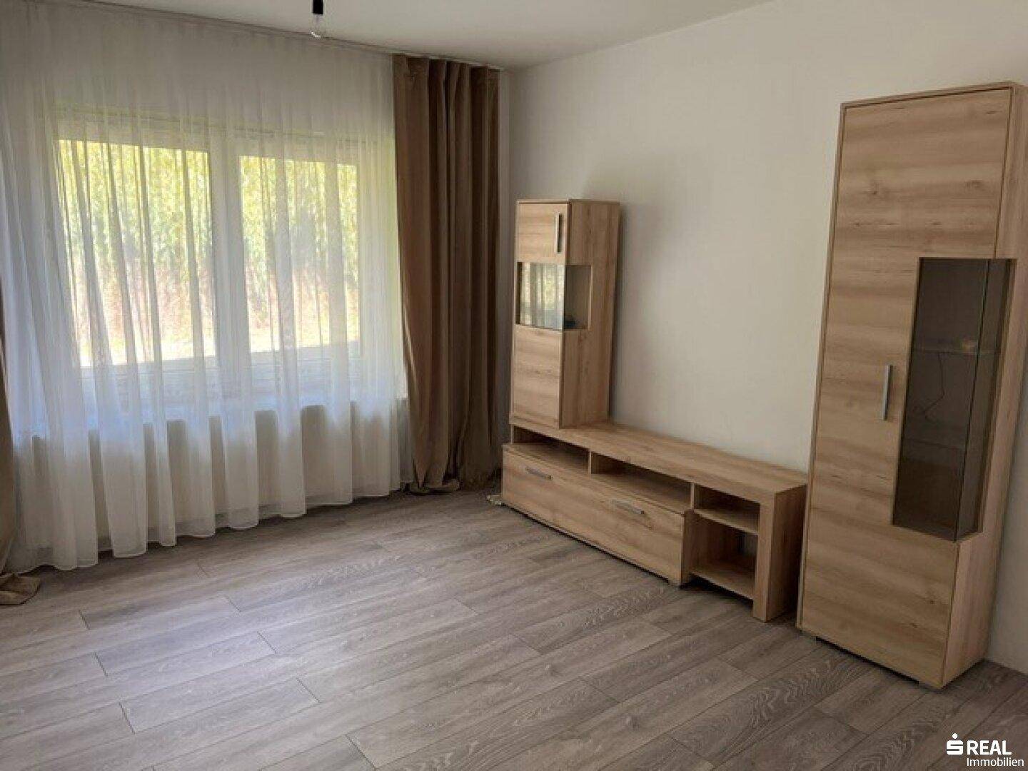 Heller Raum mit Holzmöbeln, Laminatboden und großen Fenstern mit Vorhängen.