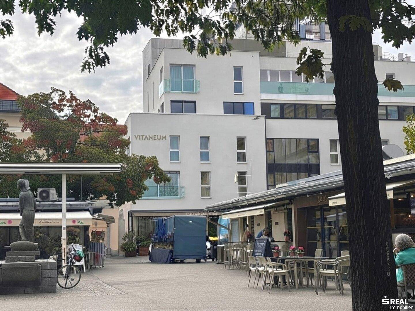 Gebäudeansicht des VITANEUM mit umliegenden Cafés und Bäumen in urbaner Umgebung.