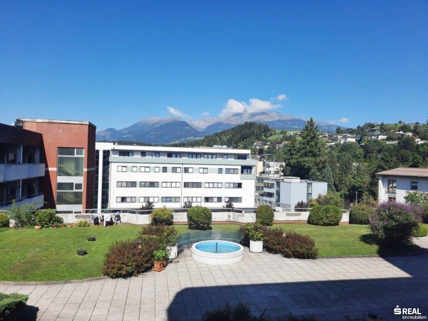 Gepflegte Außenanlage mit Rasenfläche, Brunnen und beeindruckendem Bergblick im Hintergrund.