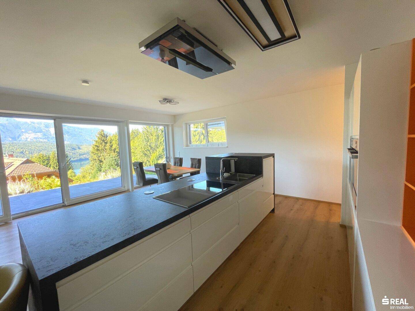 Moderne Küche mit Kochinsel, dunkler Arbeitsplatte und Panoramafenstern mit Seeblick.