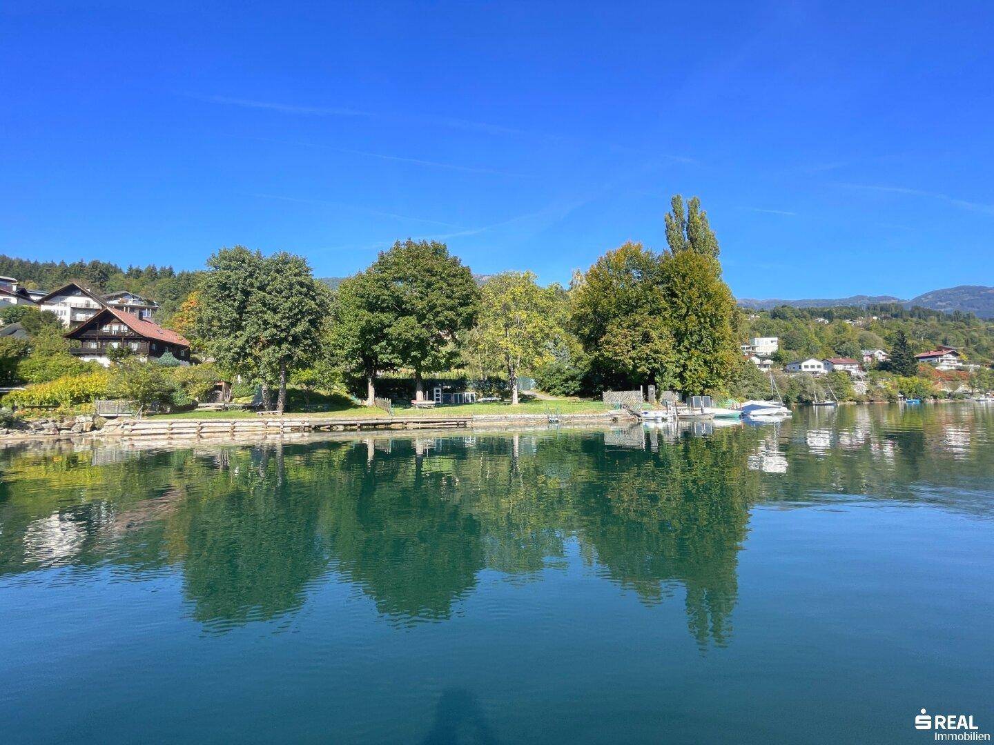 Malerischer Blick auf den See mit bewaldeten Ufern und Bergen im Hintergrund unter blauem Himmel.